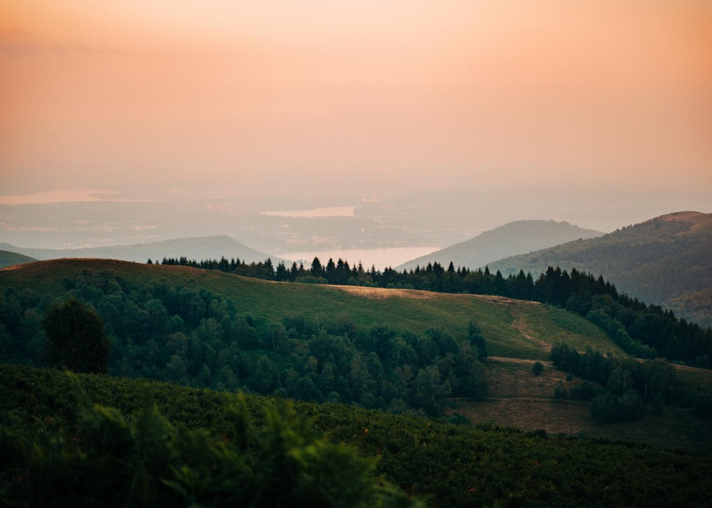 Rolling hills with large trees throughout with multiple lakes in the background and a hazy, orange sky above.