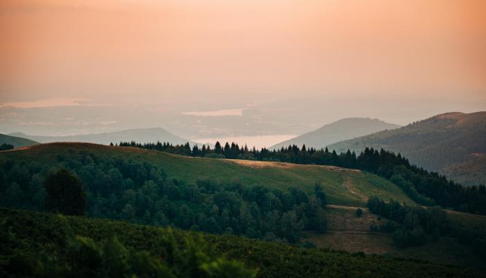 Rolling hills with large trees throughout with multiple lakes in the background and a hazy, orange sky above.