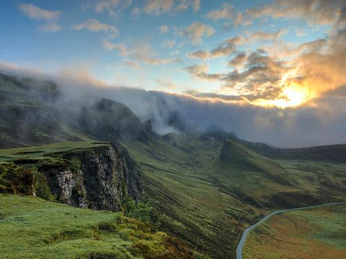 A grassy cliff overlooking a road on the right side of the image, with a cloud sunset in the background.